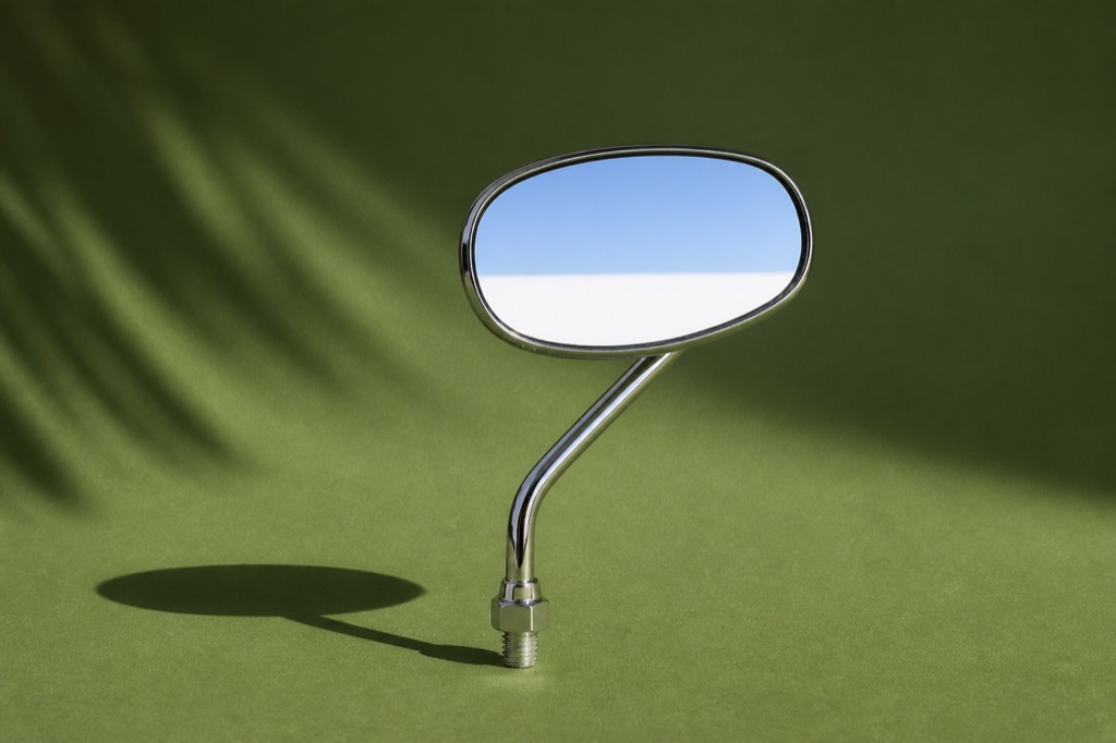 Um único retrovisor cromado de moto sobre fundo verde-palmeira, refletindo uma faixa de céu claro
